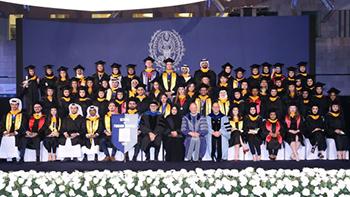 A group of more than 60 graduating seniors stand on stage with faculty members dressed in caps and gowns.